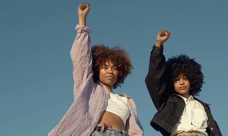 Two young black women raising their fists against a blue sky.