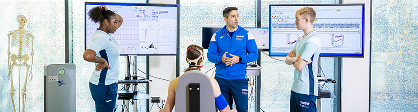 man sitting in a chair with heart monitoring equipment attached to him linked to computers and doctors standing around him