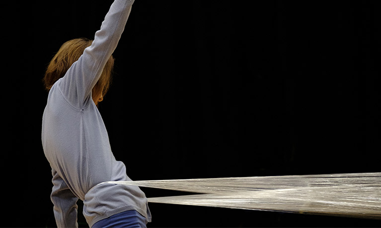 A woman facing right reaches upward with her right hand with strings wrapped around waist pulling her to the right.