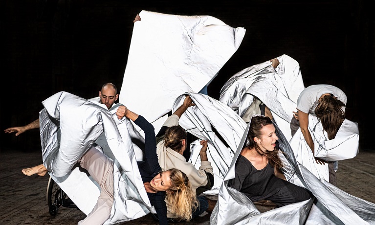Several dancers moving with white fabric in front of a black background