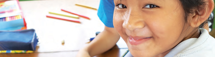 Young schoolgirl smiling at camera with coloured pencils on the table