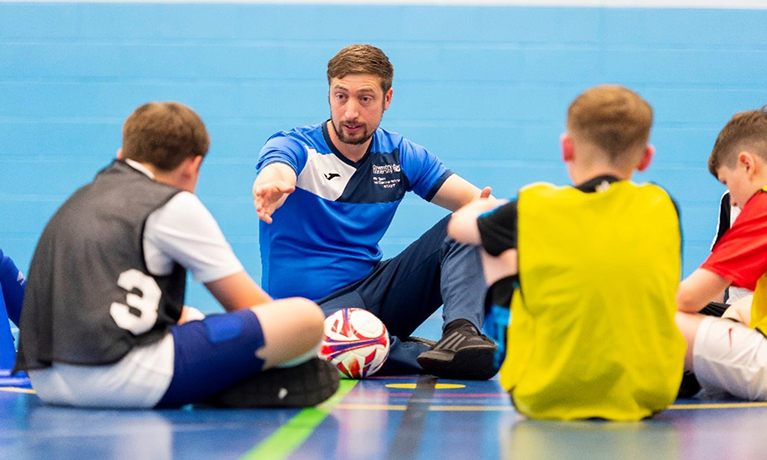 A football coach sat on the floor speaking to a group of young boys in football kits