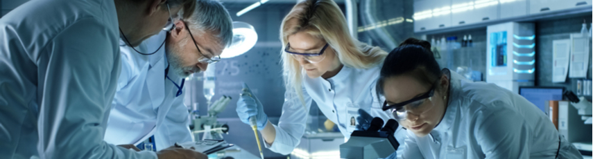 two men and two women in lab coats lean over a lab bench looking at microscopes and petri dishes.