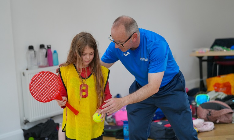 A member of Coventry University staff wearing a blue t-shirt helps a young girl with a tennis ball and racquet