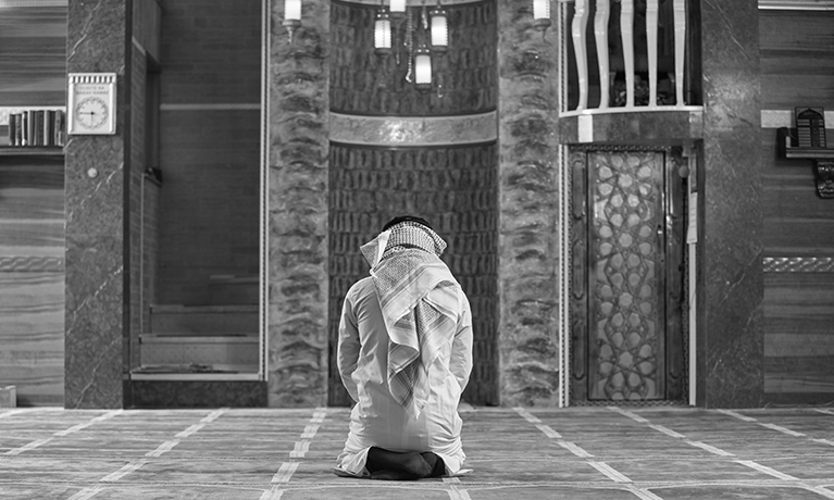 Black and white photo of the back of a man kneeling in a mosque