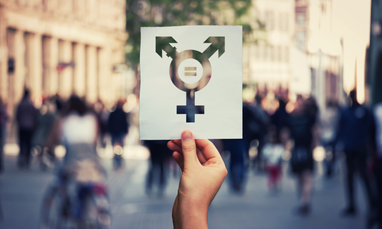 Hand holding a paper sheet with transgender symbol and equal sign inside. Equality between genders concept over a crowded city street background.