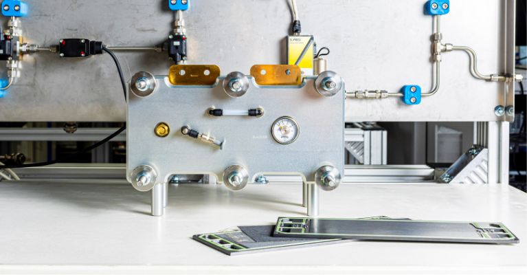 A silver hydrogen fuel cell on a table with silver piping connected to a silver wall in a lab