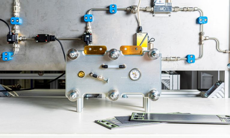 A silver hydrogen fuel cell on a table with silver piping connected to a silver wall in a lab
