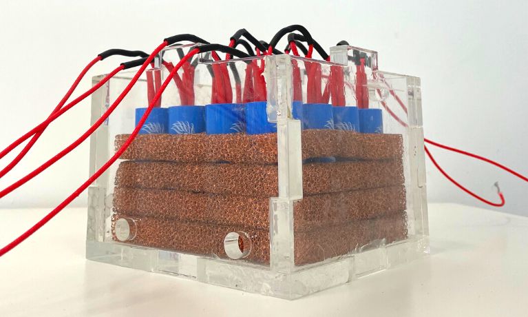 A close up of a Perspex box with 4 layers of copper coil, blue batteries and red cables 