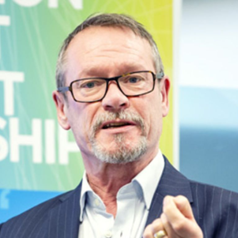 Picture of Trevor Toman wearing a suit and speaking. He's standing in front of a blurred green wall