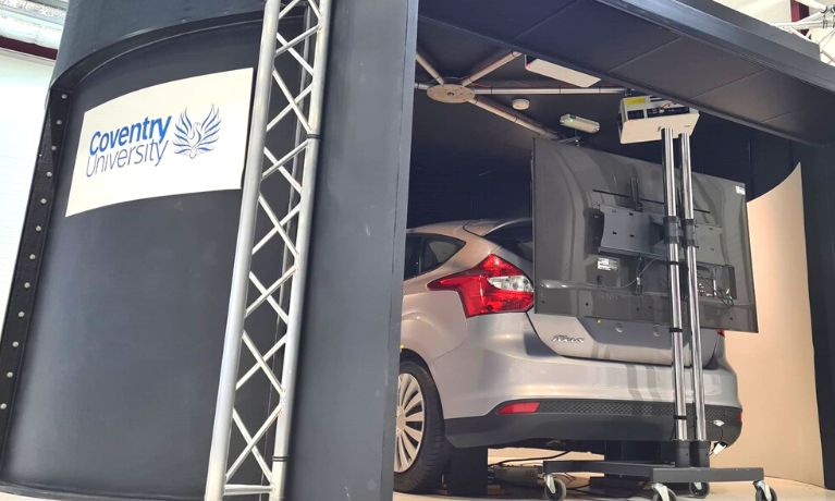 The photo shows a car inside a driving simulator. The car is silver and the simulator exterior is black with a Coventry University logo The photo shows a car inside a driving simulator. The car is silver and the simulator exterior is black with a Coventry University logo