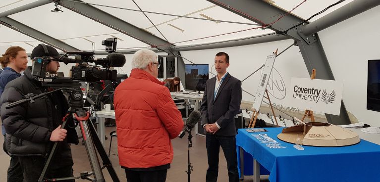 A Coventry University professor in a dark suit stands next to a blue table being interviewed by a professional film crew 