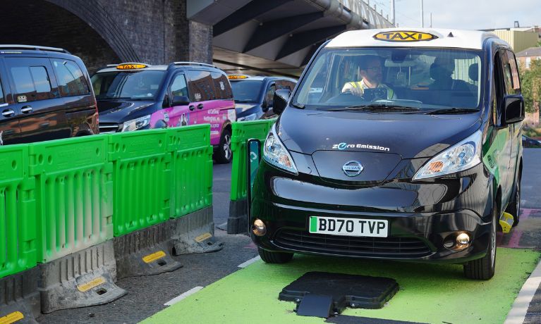 Photo of a black electric taxi with green WiCET branding on the side. The taxi is on top of a black charging pad at the taxi rank, surrounded by a green strip of tarmac on the