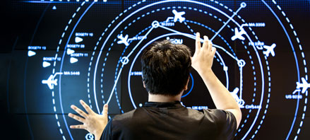 over the head photo of person with black shirt and black hair touching a huge, interactive screen with airplane icons 