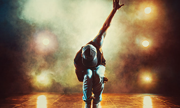 Male hip-hop dancer wearing a cap in a crouched position with lights in the background.