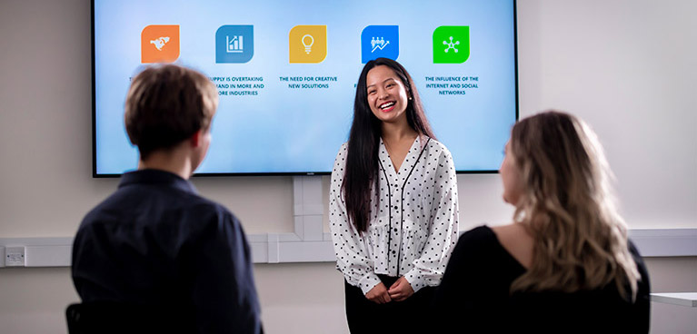smiling female student presenting in front of whiteboard to a male and female student