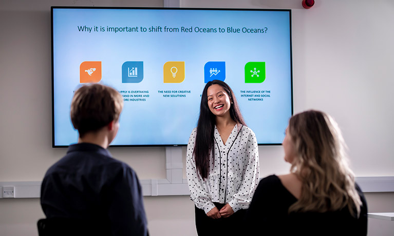 Female student stood in front of a digital screen, presenting to two students