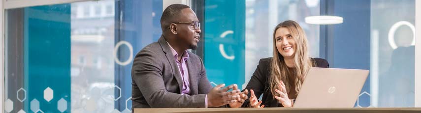 Male and female staff member chat at desk with laptop.