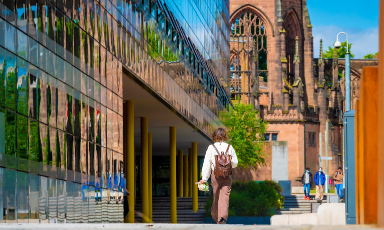 Student walking past The Hub with Coventry Cathedral ruins in the background.