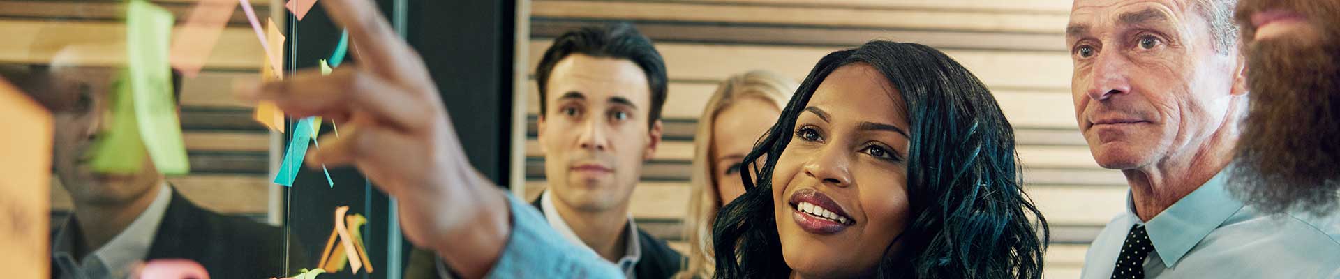 Professional woman pointing to a large screen with post notes. A group of men are looking on.