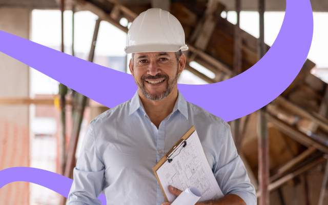 A smiling construction site manager wearing a hard hat holds a clipboard with building plans inside a partially built structure.