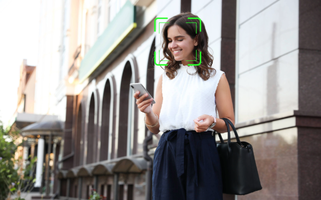 Woman walking outdoors while looking at her phone, with a green facial recognition frame around her face.