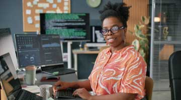 Woman wearing glasses chatting to another person with graphics on a screen in the background. Image is overlaid with a small banner saying Coming soon. 