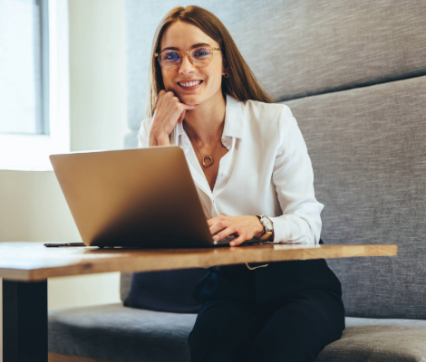 Woman sitting at a table with a laptop, smiling toward the camera.