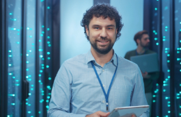 A smiling man wearing a light blue shirt and lanyard stands in a server room, holding a tablet, with rows of illuminated data servers behind him and another person working in the background.