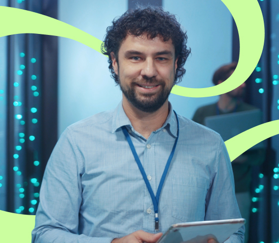 Man smiling while holding a tablet in a server room with another person working in the background.