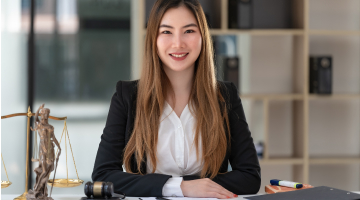 Person in business attire seated at a desk in an office, with a legal scales statue and gavel visible on the desk and shelving in the background.