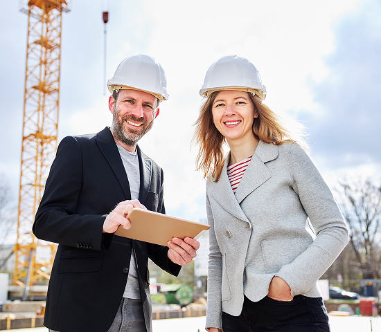 Two individuals in white hard hats at a construction site, one pointing at a tablet while the other observes, with a crane and construction materials in the background.