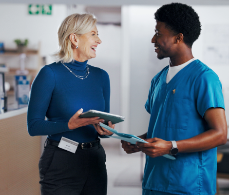 Two colleagues in a healthcare setting smiling and talking while holding tablets.