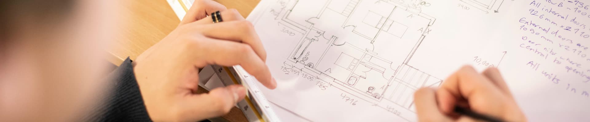 Close-up of a person using a ruler and pencil to work on detailed architectural floor plan drawings and design calculations at a desk.
