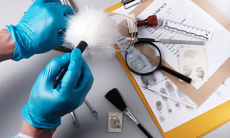 Gloved hands holding a fingerprint brush over a desk covered with forensic tools and fingerprint samples.