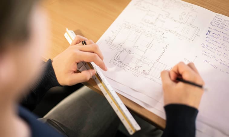 Close-up of a person using a ruler and pencil to work on detailed architectural floor plan drawings and design calculations at a desk.