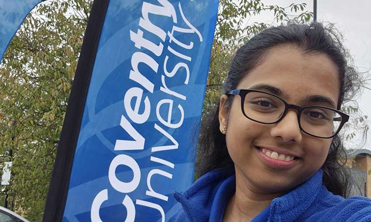 Sri outside on campus standing beside a Coventry University flag