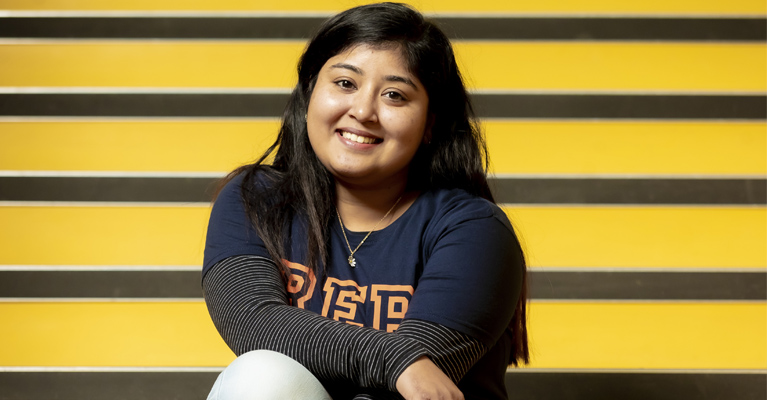 A student sits on a yellow staircase, smiling at the camera.