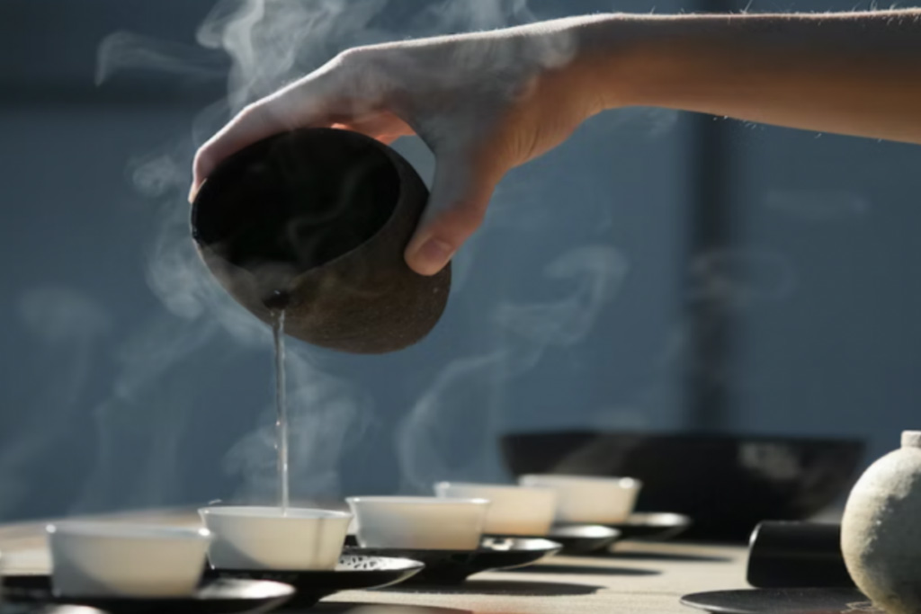 A close up of a hand pouring tea from a jug, with steam rising from the tea.