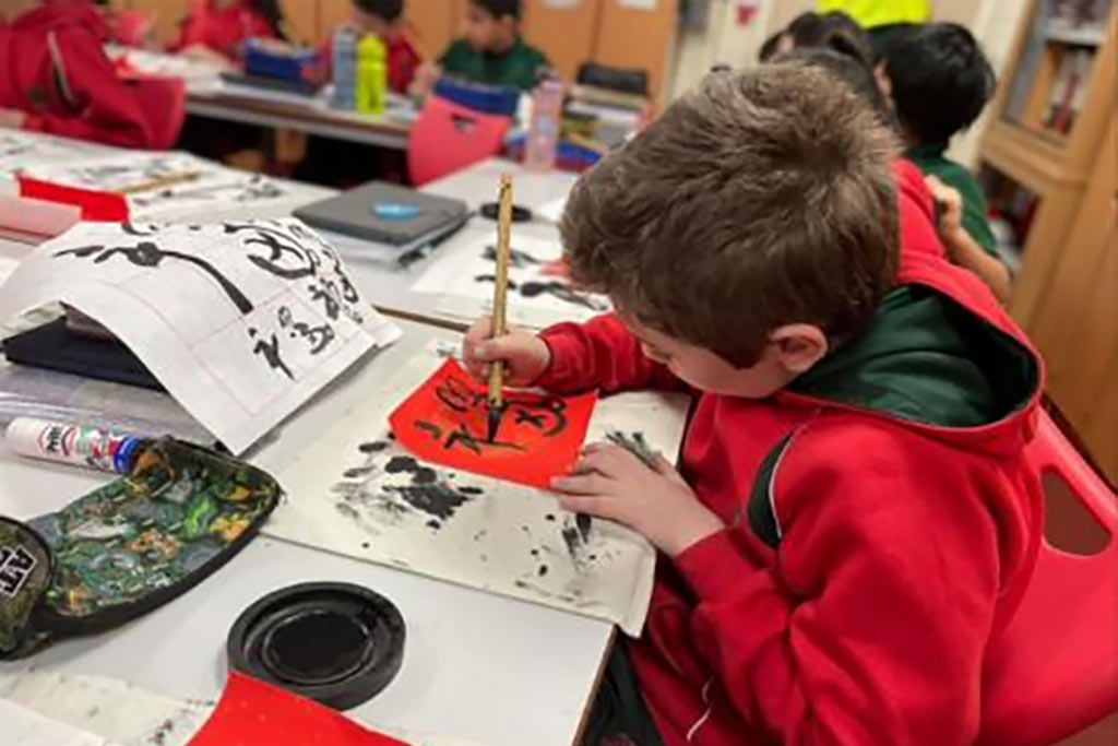 A school student sat at a table using a brush and ink to draw Chinese calligraphy characters.