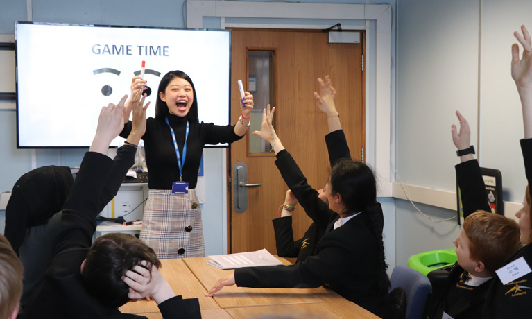A Chinese teacher smiling in front of a whiteboard in a school classroom, as students hold up their hands to answer a question.