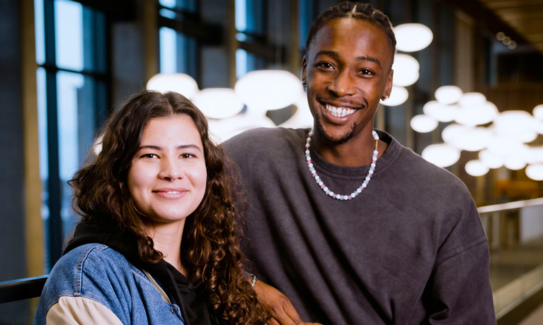 Two young, fashionably dressed students smiling at the camera with modern hanging lights in the background.