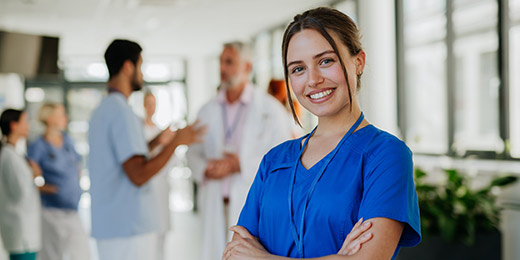 A nurse in a hospital corridor wearing blue uniform smiling and crossing arms