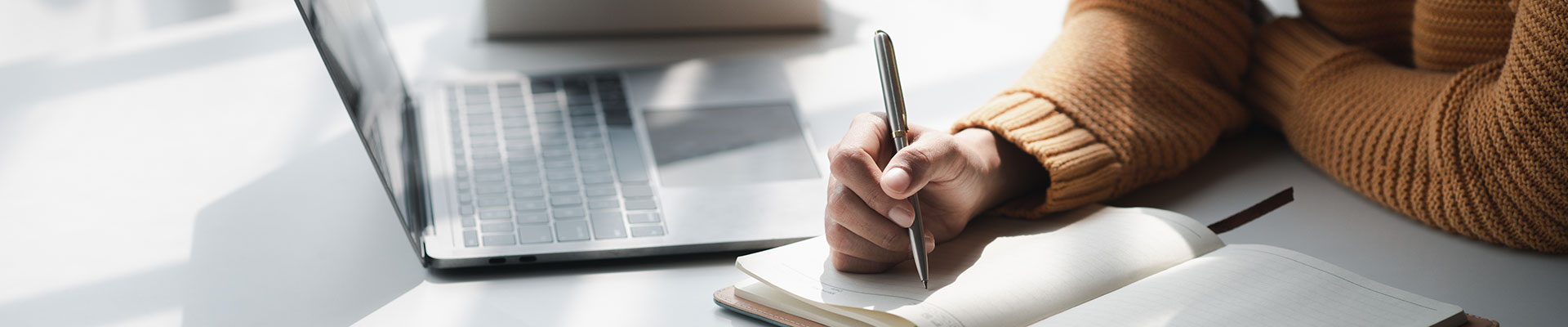 Someone writing in a notebook at a desk next to a laptop