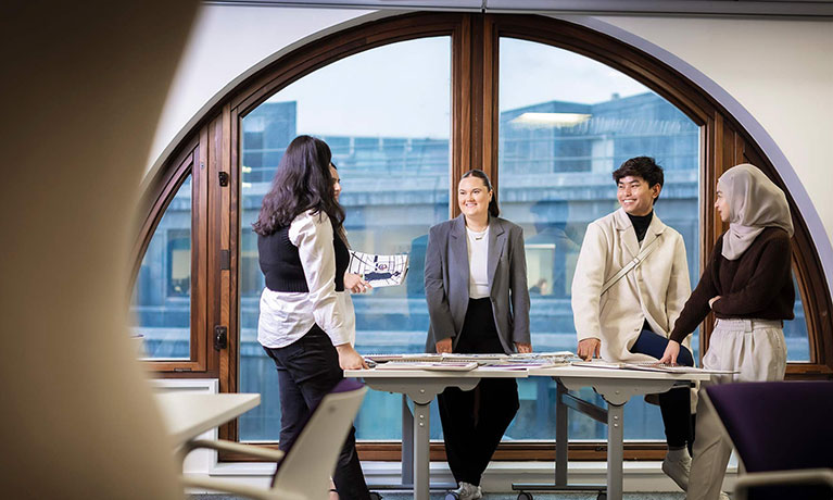 group of students standing around a table with the London skyline in the background in the window
