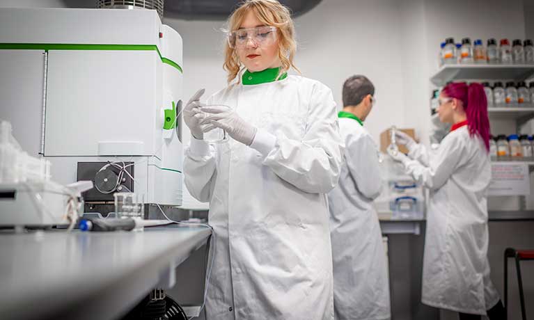 Female in a white lab coat holding a plastic beaker, with a male and female standing behind wearing lab coats