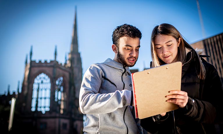 2 students looking at a clipboard with coventry cathedral ruins in the background