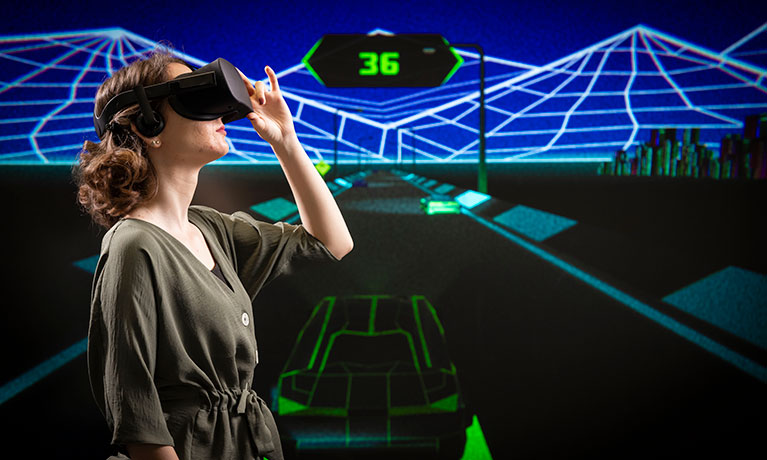 Student wearing VR goggles standing in front of digital back drop of motor car racing track 