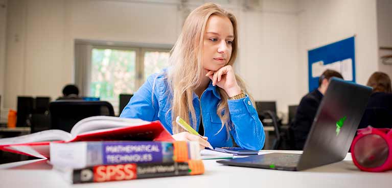 A female student sat at a desk holding a pen and looking at a laptop.