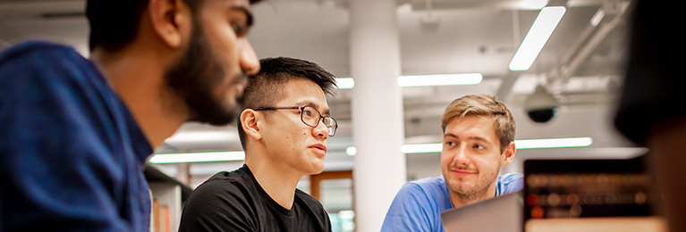 Three students chatting at a desk with laptops